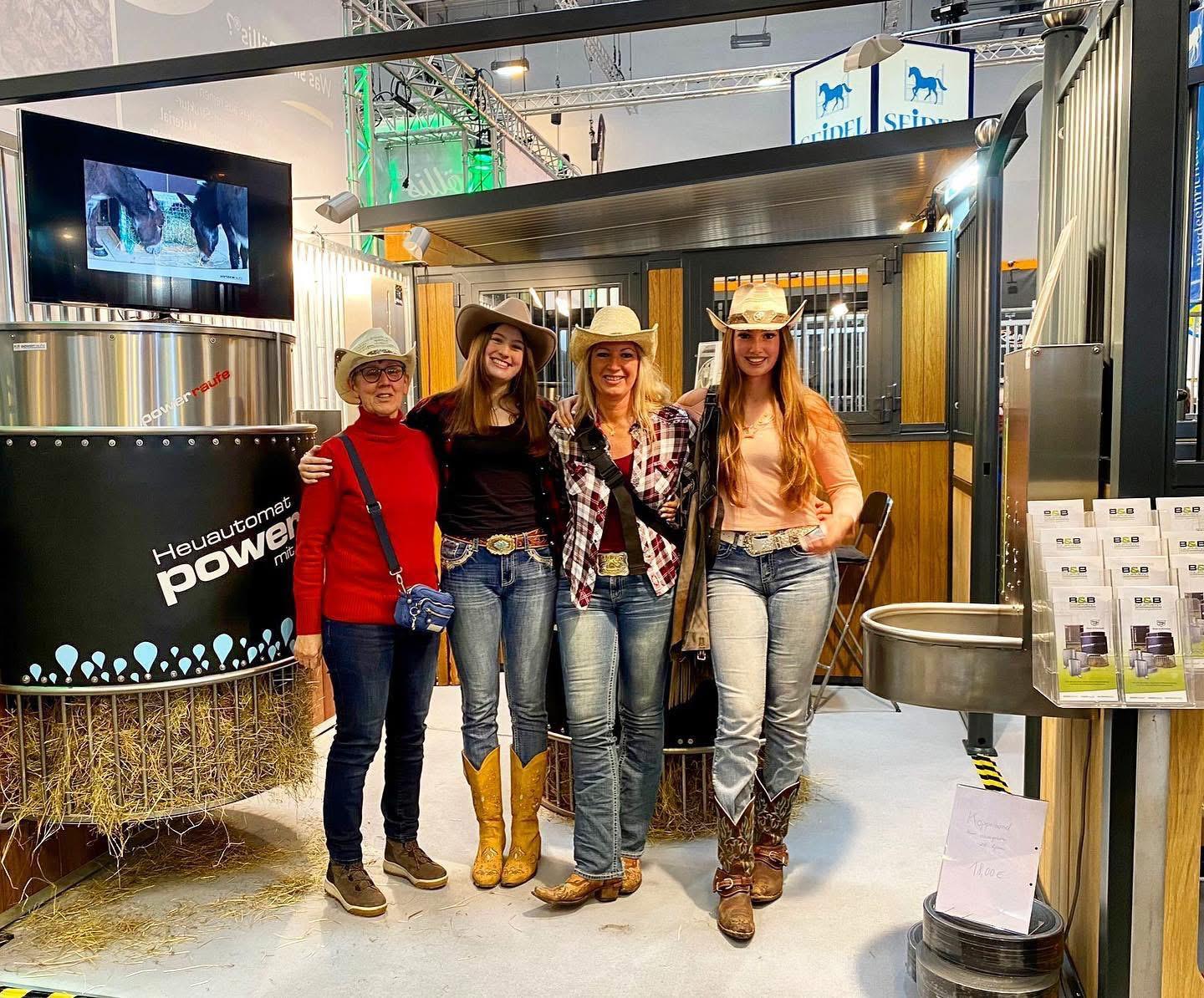 Vier Frauen in Westernkleidung stehen auf einer Pferdemesse vor einem Messestand mit zeitgesteuerter Futterautomaten wie Heuautomaten und Kraftfutterautomaten.